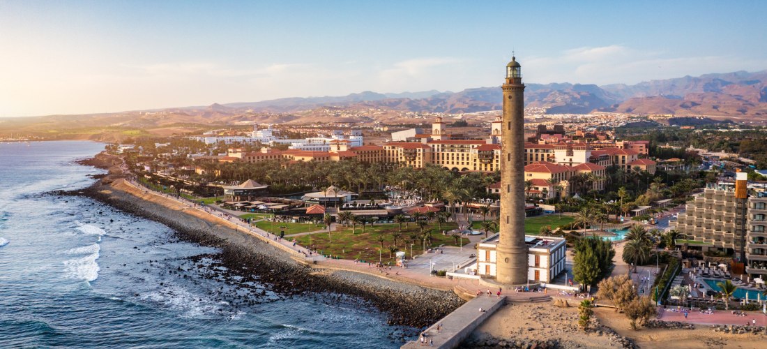 Luftaufnahme vom Leuchtturm von Maspalomas an der Küste Gran Canarias, flankiert von Promenade, Hotels und Bergen im Hintergrund.