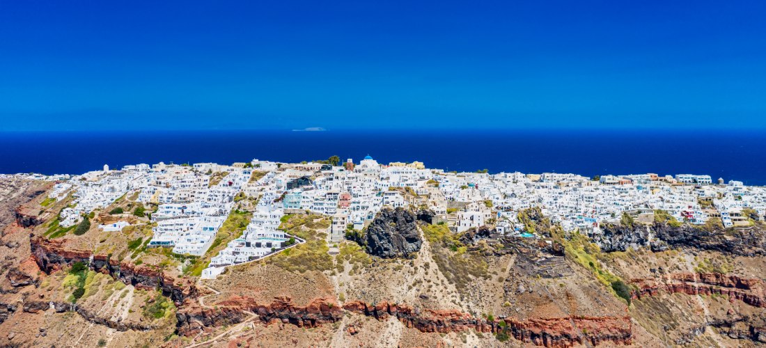 Imerovigli auf Santorini aus der Luft: weiße Häuser auf der Kraterkante, rote Lavaschichten und der Skaros-Felsen vor tiefblauer Ägäis