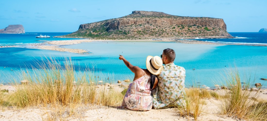 Ein Paar sitzt auf einer sandigen Düne mit Blick auf die türkisfarbene Lagune von Balos auf Kreta. Die Frau trägt einen Strohhut und zeigt mit dem Finger auf das Wasser, während der Mann neben ihr sitzt. Im Hintergrund liegen ein Boot, eine flache Landzun