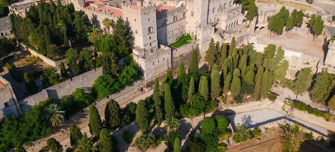 Die Luftaufnahme zeigt den mittelalterlichen Palast des Großmeisters in Rhodos-Stadt, umgeben von einer dichten Stadtmauer und üppigen Zypressenhainen. Die imposanten Mauern und Türme der Burg bestehen aus hellem Stein und verleihen dem Bauwerk ein mächti
