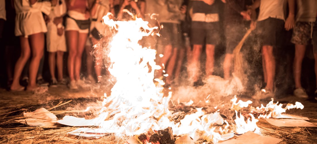 Menschen stehen nachts barfuß um ein Feuer am Strand, das auf Holzstücken und Papier brennt. Der sandige Boden und der Rauch vermitteln eine warme, feierliche Atmosphäre. Einige Zuschauer filmen mit ihren Handys.