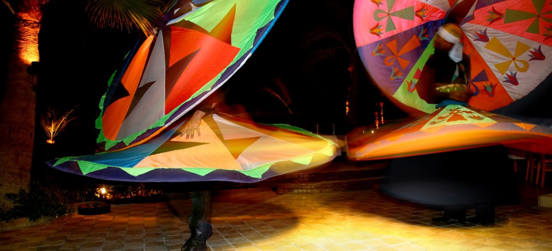 Zwei Tänzer führen bei Nacht einen traditionellen Tanoura-Tanz mit leuchtend bunten, drehenden Kostümen auf. Die Szene spielt sich auf einem beleuchteten Platz mit Palmen ab.