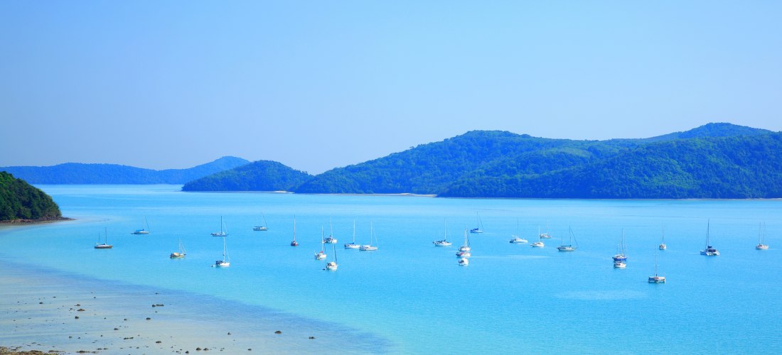 Panoramablick auf eine ruhige, türkisfarbene Bucht auf Phuket mit zahlreichen weißen Segelbooten im Wasser. Im Hintergrund erstrecken sich grün bewachsene Hügelinseln unter einem klaren, blauen Himmel. Der Sandstrand im Vordergrund wirkt naturbelassen.