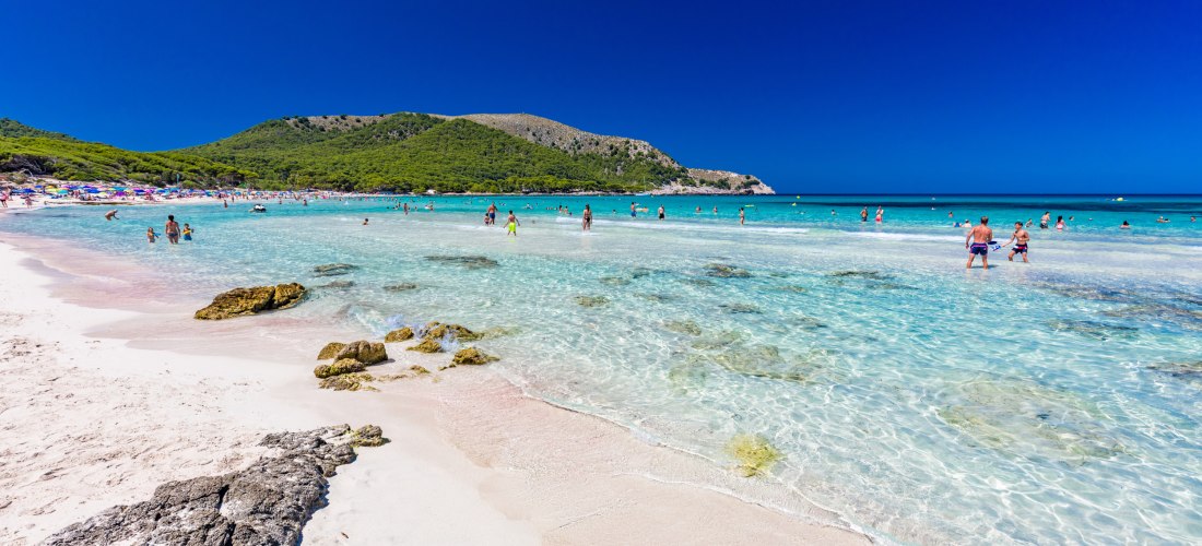 Menschen genießen das seichte, türkisfarbene Wasser am Strand Cala Agulla auf Mallorca, umgeben von grünen Hügeln und weißem Sand