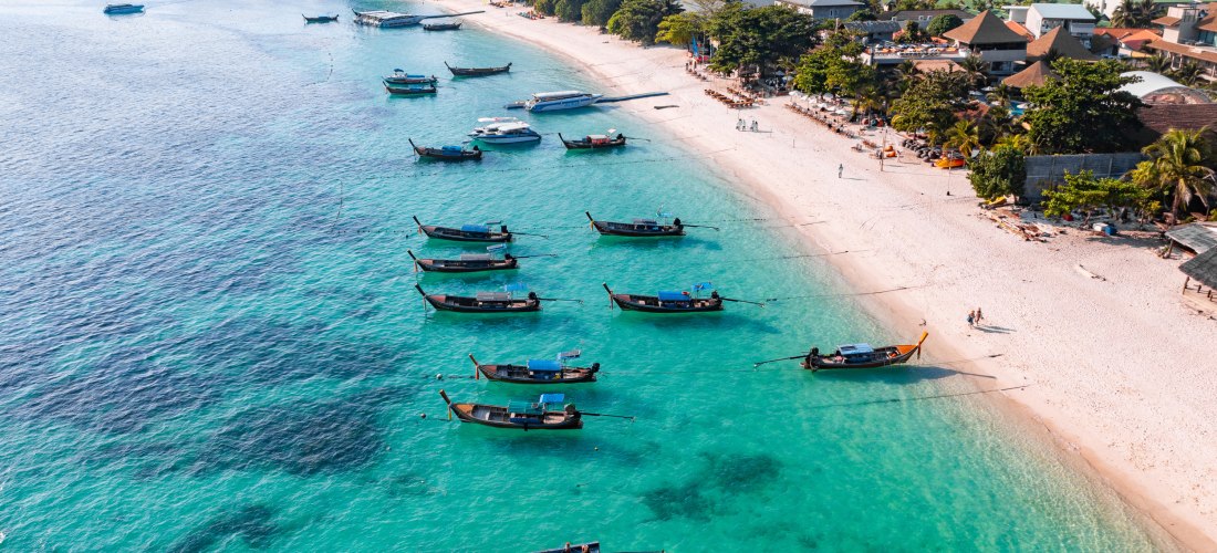 Luftaufnahme eines tropischen Strandes mit türkisblauem Wasser und traditionellen Longtail-Booten, die nahe der Küste ankern. Am weißen Sandstrand liegen gemütliche Resorts unter Palmen, im Hintergrund dichter Wald und Berge.