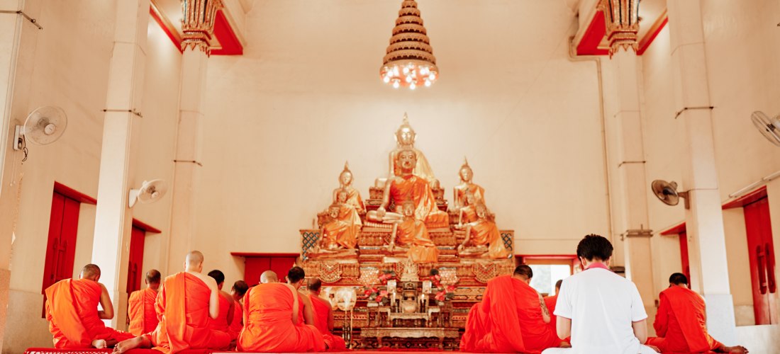 Innenraum eines buddhistischen Tempels mit goldener Buddha-Statue. Eine Gruppe von Mönchen in orangenen Roben sitzt auf dem Boden in stiller Meditation, eine weitere Person in weißer Kleidung sitzt ihnen gegenüber. Die Atmosphäre wirkt friedlich und spiri