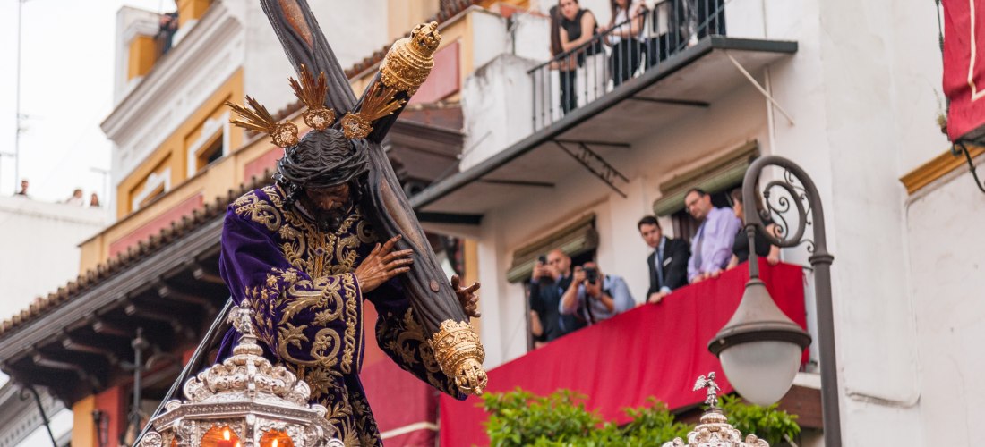 Detailaufnahme einer prachtvoll geschmückten Jesusfigur mit Dornenkrone und Kreuz während einer Karfreitagsprozession, mit Zuschauern auf Balkonen und Gebäuden im Hintergrund