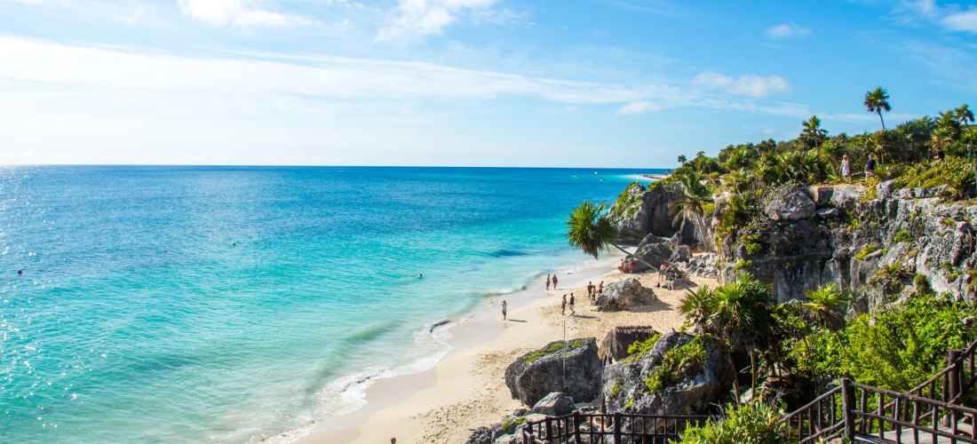 Wunderschöner Sandstrand mit türkisblauem Meer, umgeben von Felsen und tropischer Vegetation. Menschen spazieren und entspannen am Strand unter einem sonnigen, blauen Himmel