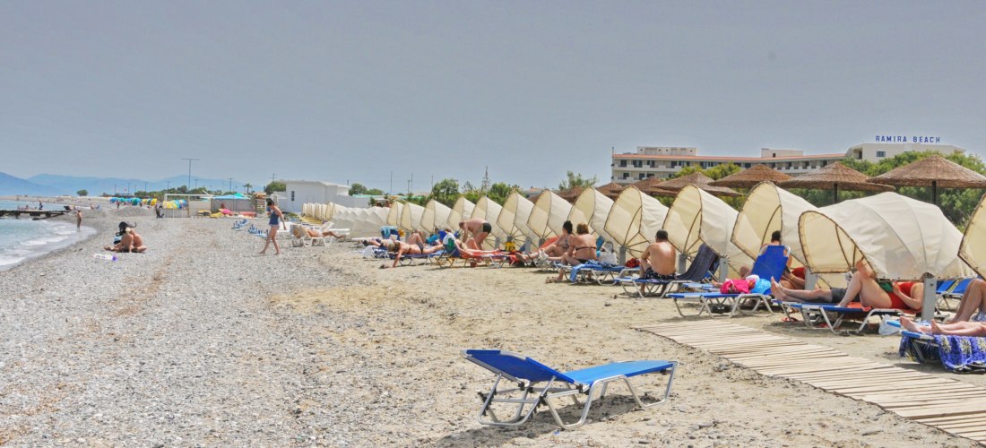 Strandabschnitt mit Kies und Sand nahe Kos-Stadt. Zahlreiche Liegen mit halbrunden Sonnenschirmen stehen in einer Reihe, dahinter sieht man Hotelgebäude mit dem Schriftzug 'Ramira Beach'. Menschen sonnen sich und genießen das Meer.