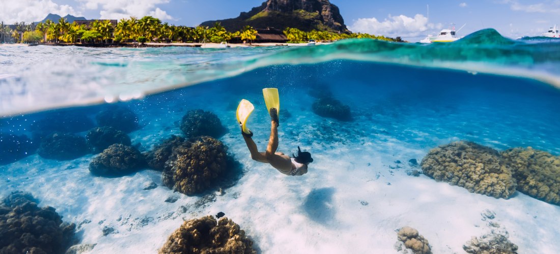 Dieses atemberaubende Bild zeigt eine faszinierende Kombination aus Über- und Unterwasserwelt. Im unteren Teil des Bildes taucht eine Person mit gelben Flossen und Schnorchel in das kristallklare, blaue Wasser ein, umgeben von weichen Sandböden und Korall