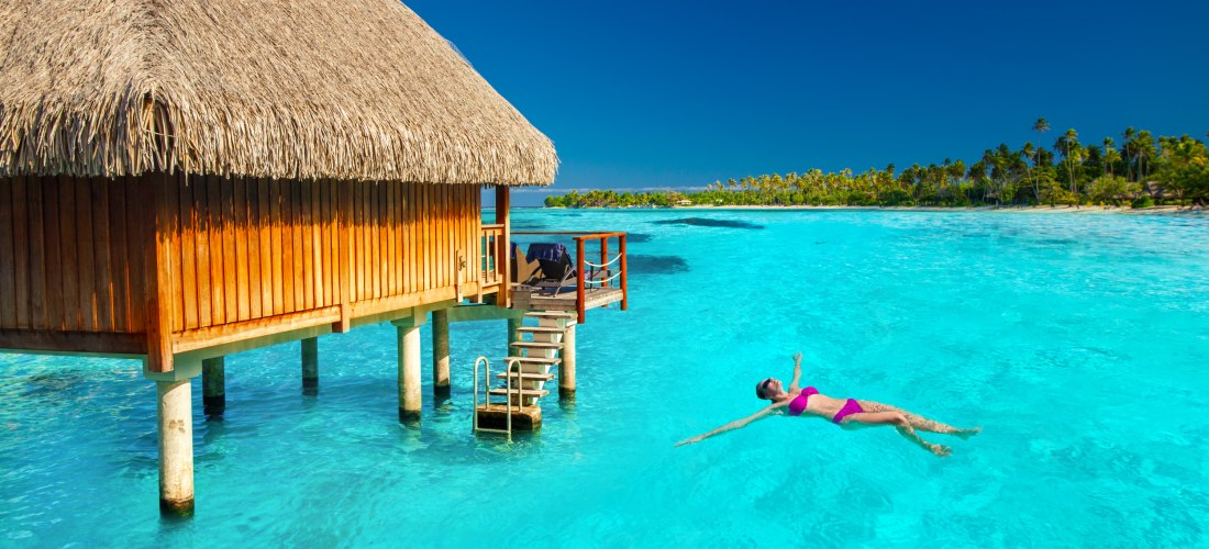 Eine Frau in pinkem Bikini treibt entspannt auf dem türkisblauen Wasser vor einem Wasserbungalow mit Strohdach. Im Hintergrund Palmeninsel, strahlend blauer Himmel und tropische Idylle. Die Szene vermittelt Ruhe und pure Entspannung in der Südsee.