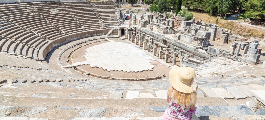 Eine Frau mit Sonnenhut und Sommerkleidung sitzt in den oberen Rängen eines antiken Amphitheaters in Ephesos, Türkei. Sie blickt auf die halbkreisförmige Bühne mit umliegenden Ruinen und grüner Landschaft im Hintergrund.