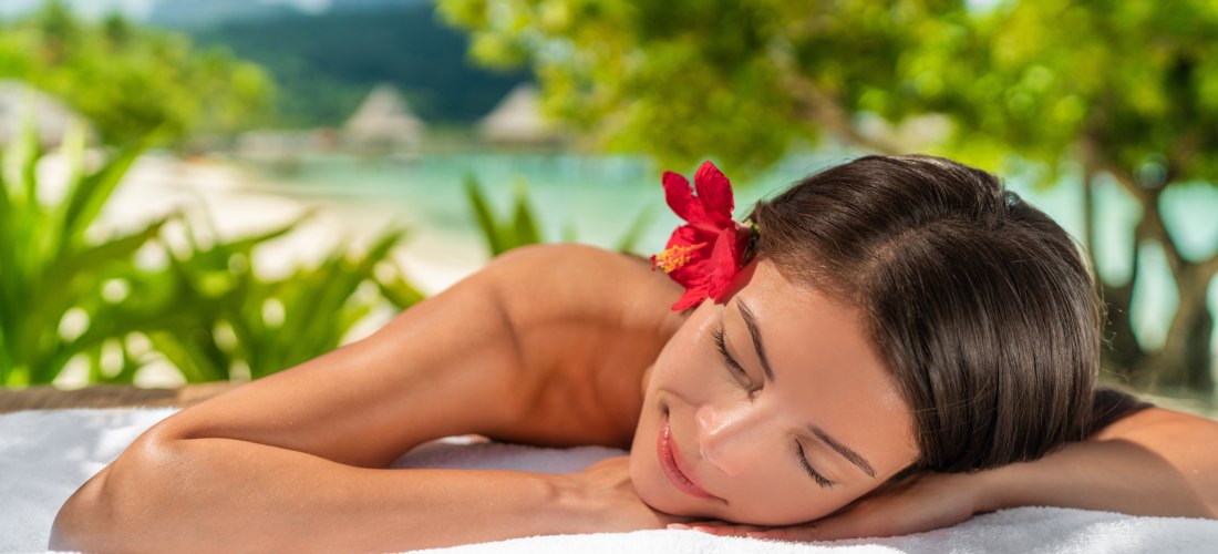 Frau liegt entspannt auf einem weißen Handtuch im Spa-Bereich eines tropischen Resorts. Sie hat die Augen geschlossen und eine rote Hibiskusblüte im Haar. Im Hintergrund sind grüne Pflanzen, Palmen und verschwommen das Meer sowie Überwasserbungalows zu se