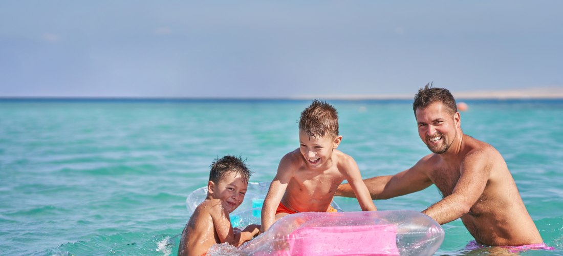 Ein Vater spielt mit zwei kleinen Jungen lachend im türkisfarbenen Meer. Die Kinder sitzen auf einer aufblasbaren pinken Luftmatratze. Im Hintergrund ist der weite Ozean zu sehen, die Stimmung ist fröhlich und unbeschwert.