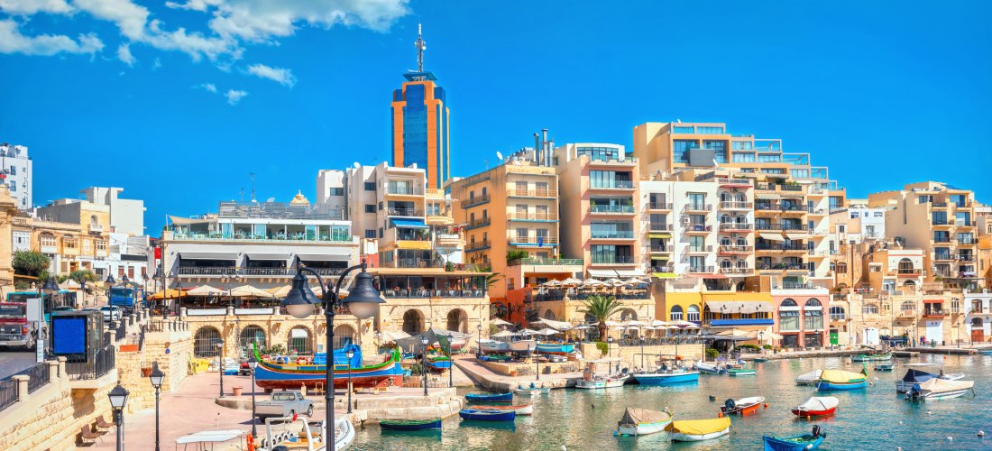 Spinola Bay in St. Julian’s auf Malta mit bunten Fischerbooten, modernen Gebäuden und Restaurants am Wasser