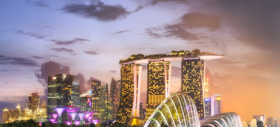 Blick auf die nächtliche Skyline von Singapur mit dem ikonischen Marina Bay Sands Hotel, das mit seiner markanten Dachplattform über drei Türmen beleuchtet ist. Im Vordergrund leuchten die futuristischen Gewächshäuser der Gardens by the Bay in sanften Far