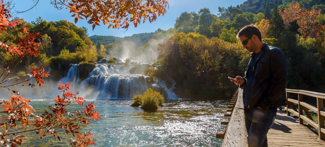 Ein Mann mit Sonnenbrille und schwarzer Lederjacke steht entspannt auf einem hölzernen Steg und blickt auf sein Smartphone. Im Hintergrund rauscht ein mehrstufiger Wasserfall durch eine von herbstlich gefärbten Bäumen gesäumte Landschaft. Die Sonne schein