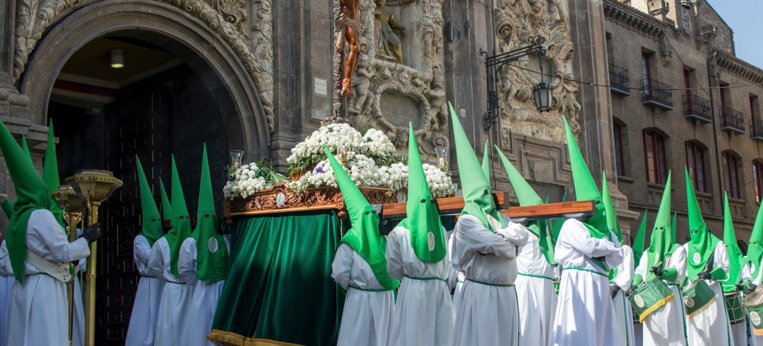 Religiöse Prozession vor einer kunstvoll verzierten Kirche, bei der Männer in weißen Gewändern und grünen Spitzhauben eine Statue von Jesus am Kreuz auf einem blumengeschmückten Tragegestell tragen