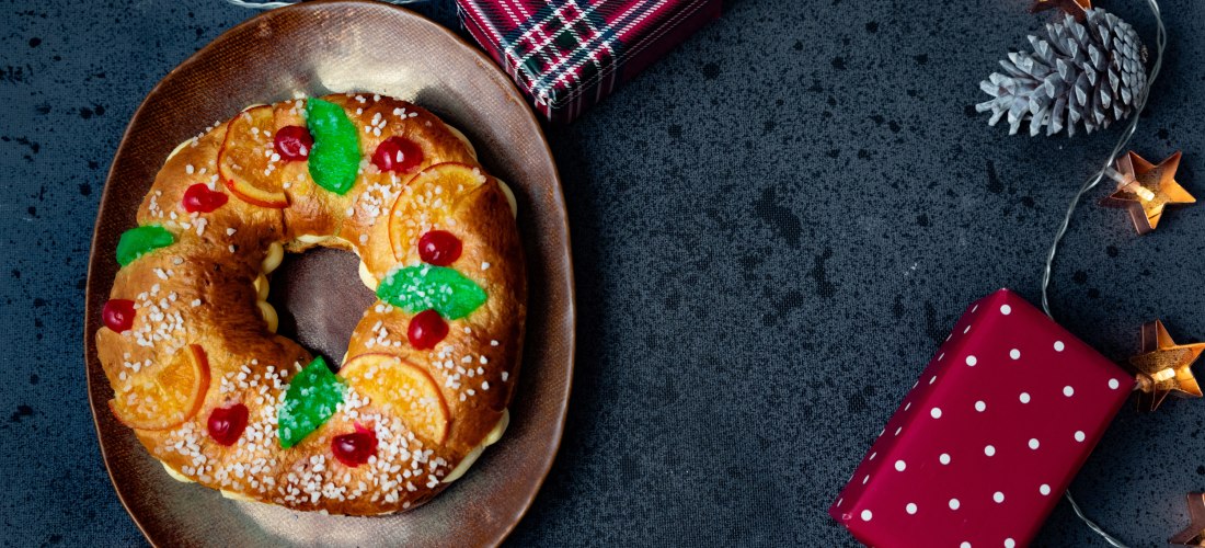 Ein traditioneller spanischer Weihnachtskuchen in Kranzform liegt auf einem braunen Teller. Der Kuchen ist mit kandierten Früchten, Zucker und Orangenscheiben dekoriert. Daneben stehen zwei bunt verpackte Geschenke. Die Szenerie ist von Tannenzapfen, Ster