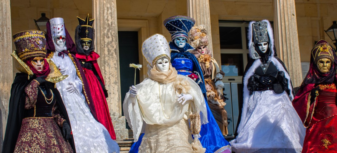 Karneval auf Korfu: Eine Gruppe kunstvoll maskierter Menschen steht in prächtigen Gewändern vor einem neoklassizistischen Gebäude. Die aufwendigen Kostüme erinnern an den venezianischen Karneval und unterstreichen die kulturelle Verbindung der Insel mit I