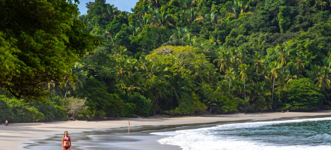 Frau spaziert am ruhigen tropischen Sandstrand entlang, umgeben von üppigem grünen Regenwald