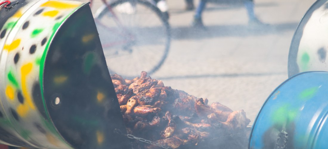 Ein großer, improvisierter Grill aus einem aufgeschnittenen Metallfass, bemalt mit bunten Farbtupfern in Grün, Gelb und Schwarz. Darauf wird eine große Menge gegrilltes Fleisch, vermutlich Jerk Chicken, über Holzkohle gegart. Rauch steigt auf. Im Hintergr