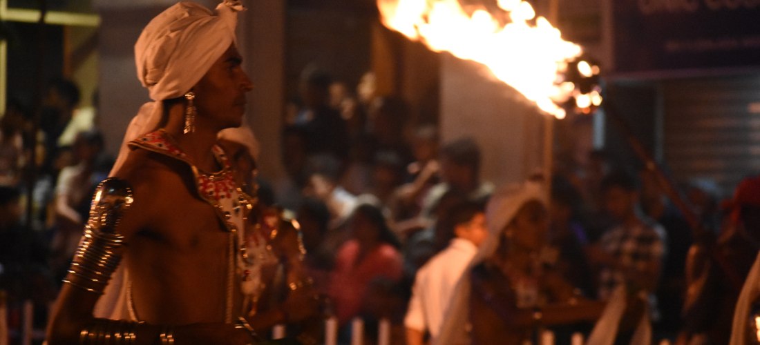Ein traditionell gekleideter Mann trägt bei Nacht eine große, brennende Fackel während einer Prozession. Er trägt weiße Kleidung mit Schmuckelementen und eine Kopfbedeckung. Im Hintergrund sind Zuschauer und weitere Teilnehmer der Parade zu sehen.