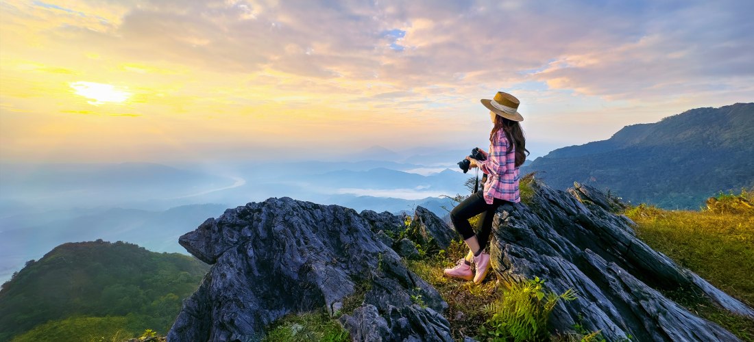 Eine Frau mit Kamera sitzt auf einem Felsen und blickt beim Sonnenaufgang über eine weite Berglandschaft mit Nebel in den Tälern. Sie trägt Wanderschuhe, Strohhut und kariertes Hemd.