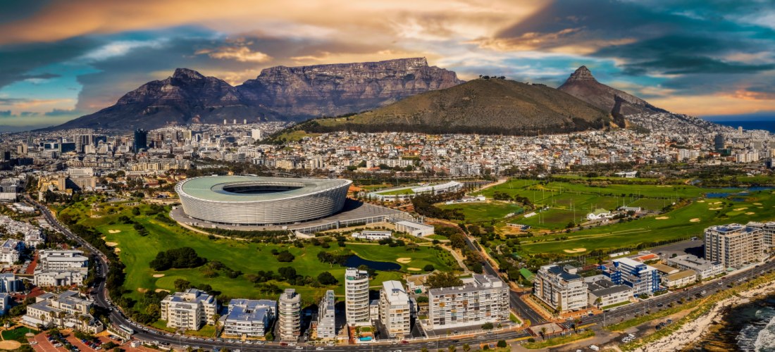 Panoramaaufnahme von Kapstadt in Südafrika mit dem markanten Tafelberg im Hintergrund. Im Vordergrund ist das Cape Town Stadium gut erkennbar, daneben weitläufige Grünflächen, die Innenstadt und die Waterfront mit dem Hafen. Rechts im Bild verläuft die Kü