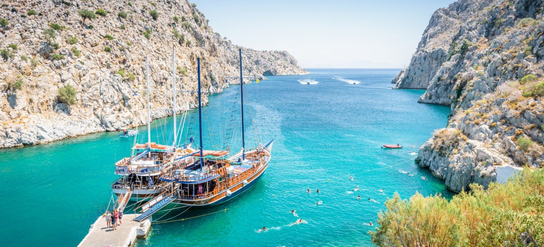 Zwei traditionelle Ausflugsboote liegen an einem Steg in einer türkisfarbenen Bucht, eingerahmt von hellen Felsen mit karger Vegetation. Menschen schwimmen im klaren Wasser, andere stehen am Ufer oder Steg. Im Hintergrund öffnet sich das Meer zwischen den