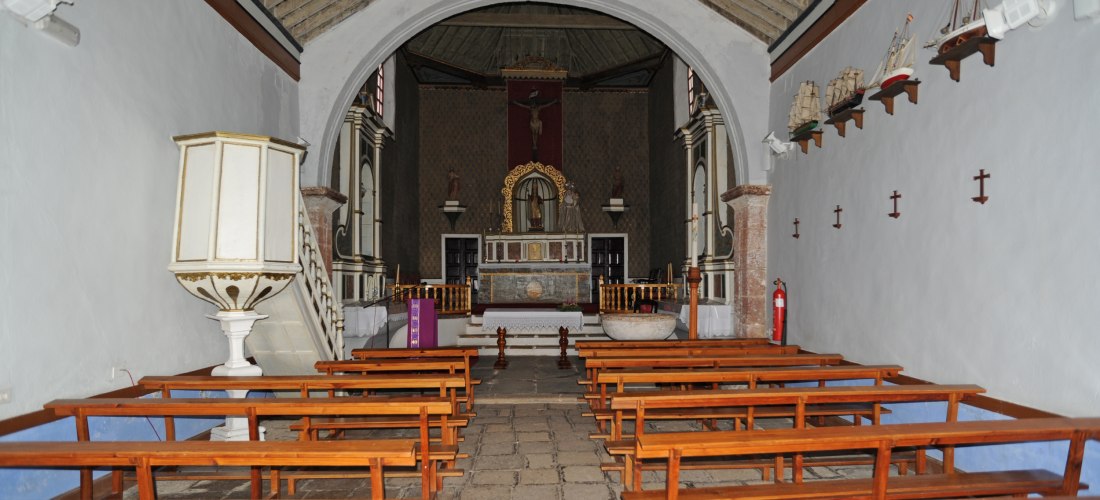 Innenraum einer kleinen Kirche mit Holzbänken, Kanzel, Altar und Kreuzigungsdarstellung. An der rechten Wand hängen Modelle von Segelschiffen auf kleinen Regalen.
