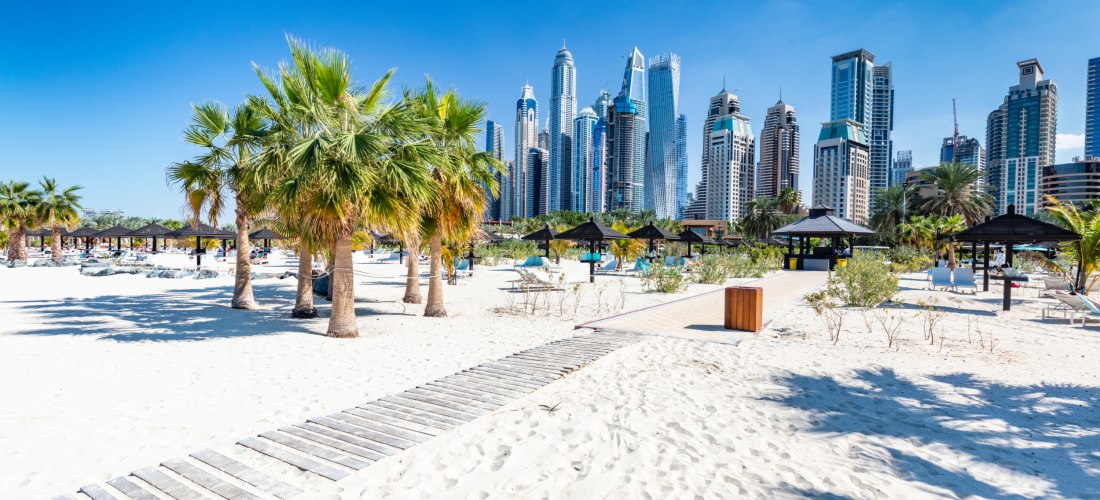 Heller Sandstrand in Dubai mit Palmen, Sonnenliegen und schattigen Pavillons. Im Hintergrund erhebt sich die moderne Skyline von Dubai Marina mit zahlreichen futuristischen Hochhäusern und Wolkenkratzern. Der Himmel ist strahlend blau, die Szene wirkt son