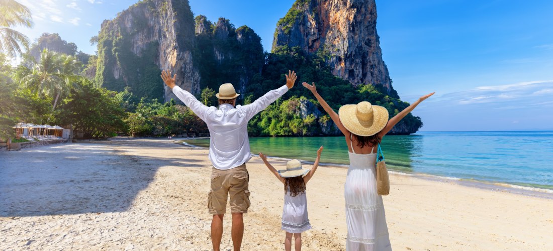 Eine Familie steht barfuß am Strand mit ausgestreckten Armen. Vor ihnen: kristallklares Wasser und beeindruckende, grüne Felsen. Alle tragen weiße Sommerkleidung und Strohhüte – Urlaubsfreude pur.