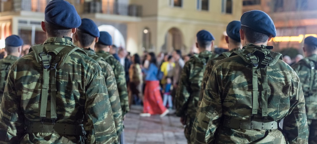 Mehrere griechische Soldaten in Tarnuniformen und blauen Baretts stehen mit dem Rücken zur Kamera bei einer offiziellen Veranstaltung oder Zeremonie. Im Hintergrund versammeln sich Zuschauer vor einem Gebäude mit Balkonen und Bögen, vermutlich auf einem ö