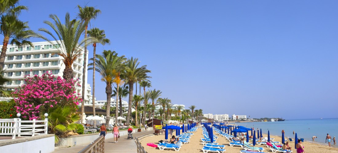 Protaras auf Zypern: palmengesäumte Strandpromenade mit Hotels sowie Reihen blauer Liegen und Sonnenschirme am breiten Sandstrand am ruhigen Meer.