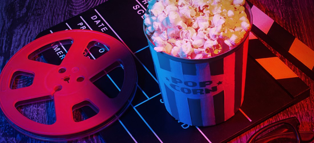 Eimer Popcorn auf einer Filmklappe, daneben eine Filmrolle und eine 3D-Brille. Das Bild ist in blau-rotem Licht gehalten und vermittelt Kinoatmosphäre.