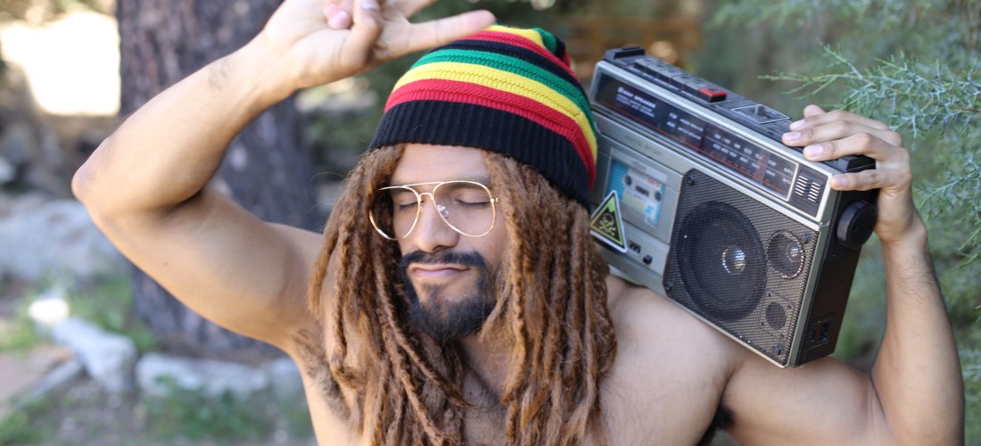 Ein junger Mann steht oberkörperfrei draußen im Grünen. Er trägt eine Mütze in den Rastafari-Farben Rot, Gelb, Grün und Schwarz sowie eine auffällige Brille. Seine Haare sind zu Dreadlocks gestylt. Auf der Schulter trägt er ein großes, altmodisches Radio.