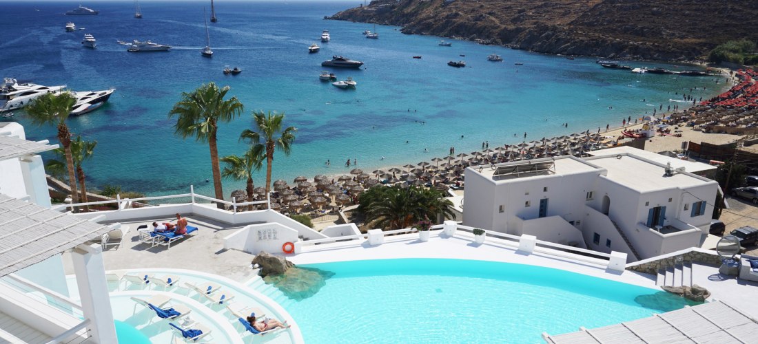 Hotelanlage mit weißem Infinity-Pool und Blick auf eine Bucht mit türkisfarbenem Wasser. Im Vordergrund Palmen und Sonnenliegen, am Strand stehen zahlreiche Sonnenschirme. Auf dem Meer ankern viele Yachten und Segelboote. Dahinter erstrecken sich felsige 