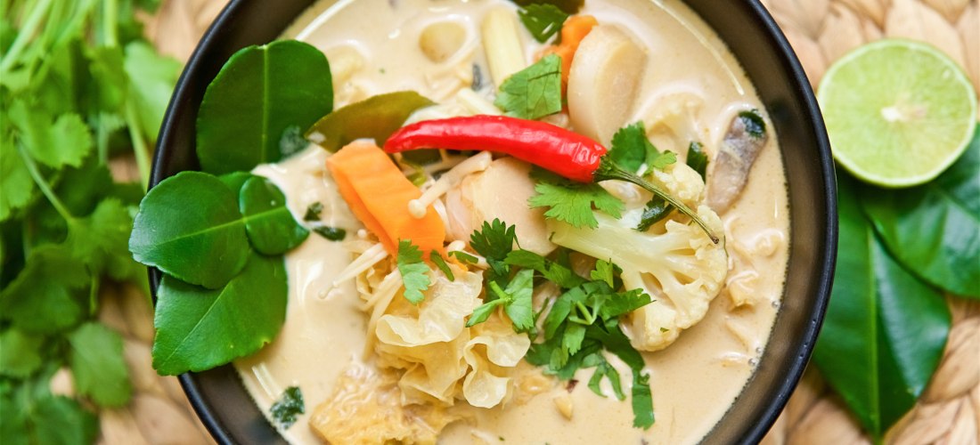 Das Bild zeigt eine schwarze Schale mit einer cremigen thailändischen Kokossuppe (vermutlich Tom Kha). Die Suppe ist mit frischen Zutaten wie Blumenkohl, Karotten, Pilzen, Koriander und einer ganzen roten Chilischote dekoriert. Um die Schüssel herum liege