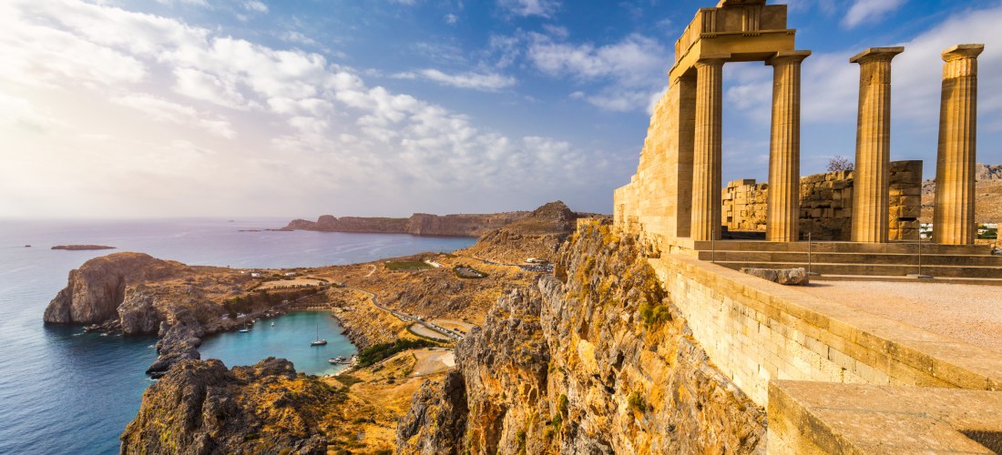 Das Bild zeigt die eindrucksvollen Ruinen der antiken Akropolis von Lindos, hoch oben auf einem steilen Felsen über dem Meer gelegen. Im Vordergrund stehen die imposanten dorischen Säulen des Athena-Tempels, die aus hellem Kalkstein errichtet sind und ein