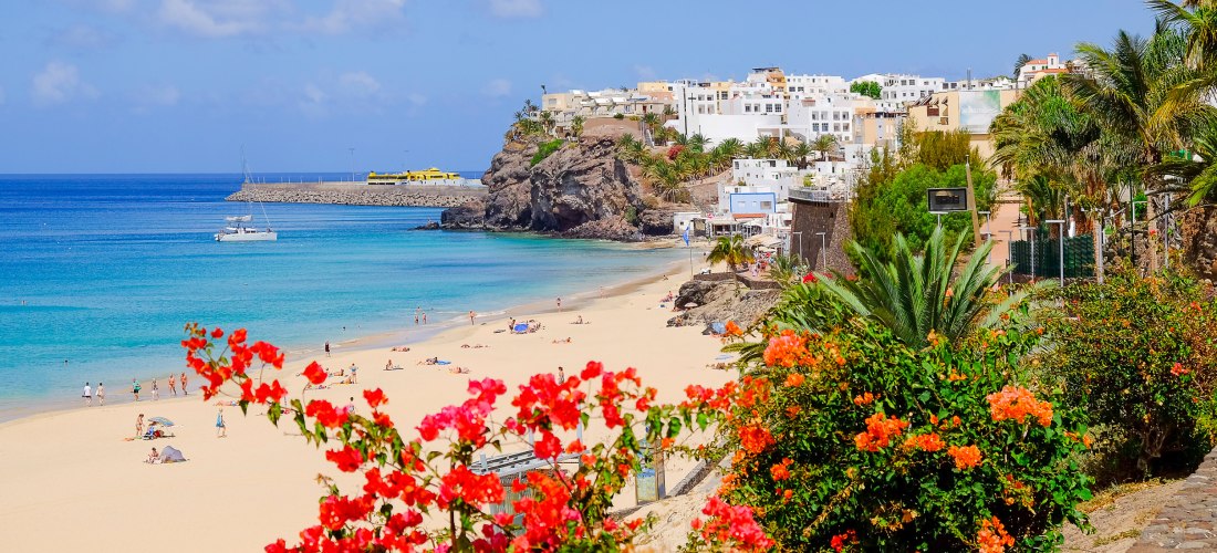 Blick auf einen hellen Sandstrand mit türkisblauem Meer. Menschen sonnen sich, bunte Blumen im Vordergrund, weiße Häuser auf einem Felsen im Hintergrund.