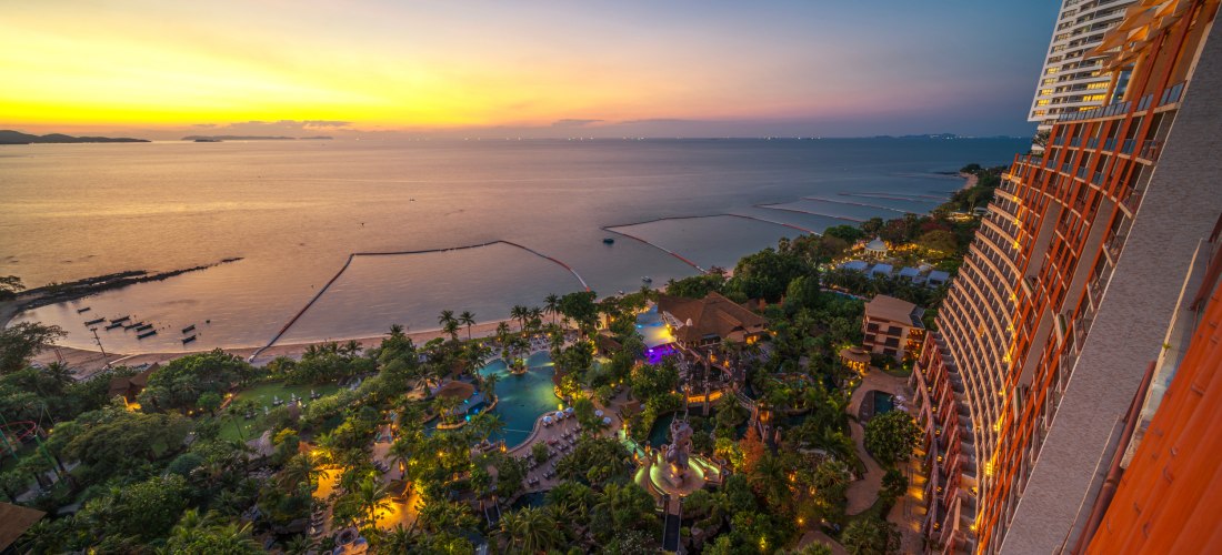 Blick vom Hochhaus auf ein beleuchtetes Luxusresort mit tropischer Gartenanlage und Pools direkt am Strand von Pattaya. Im Hintergrund ruhiges Meer bei Sonnenuntergang, der Himmel leuchtet in Gelb- und Blautönen.