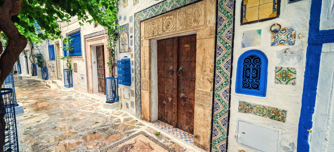 Eine mit Mosaiken verzierte Gasse in der Medina von Tunis. Links ein alter Baum, rechts eine Hausfassade mit kunstvollen Verzierungen, bunten Fliesen und einer massiven Holztür. Die Straße ist gepflastert und mit dekorativen Mosaiken versehen.