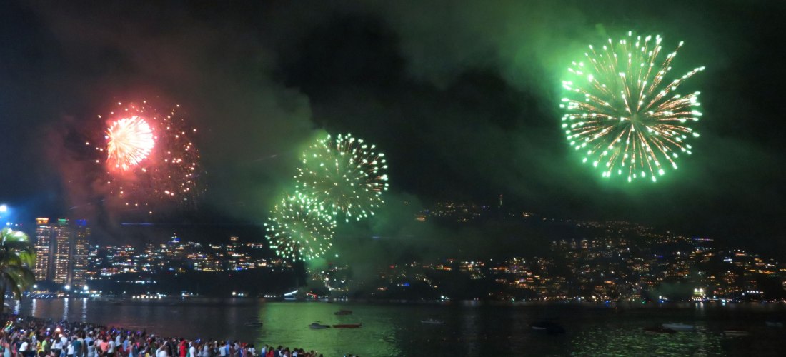 Menschenmenge am Sandstrand in Acapulco, die ein großes Feuerwerk in Grün und Rot über der Bucht bei Nacht beobachtet