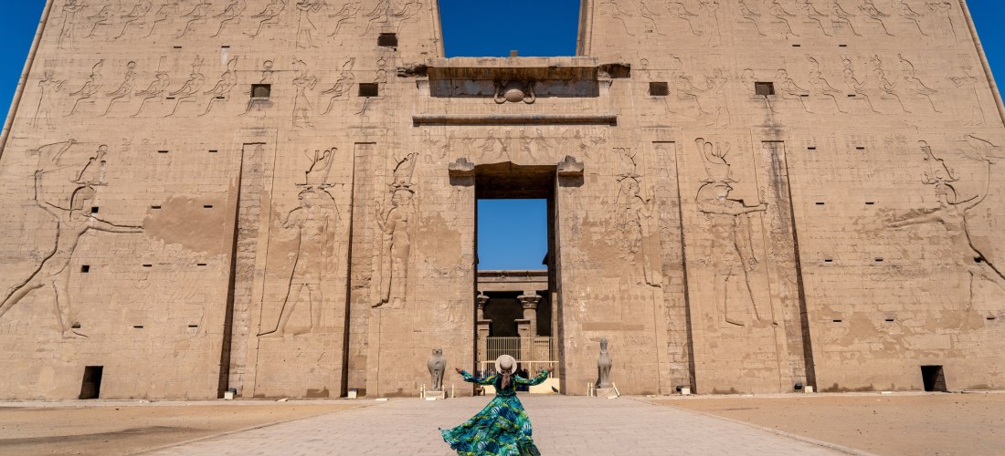 Frau in farbigem Kleid steht vor dem monumentalen Eingang eines altägyptischen Tempels mit kunstvoll gemeißelten Reliefs an der Fassade.