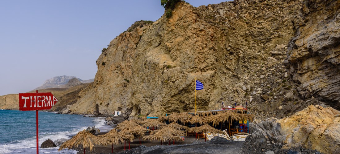 Eingang zum Strand Therma auf Kos mit Felsen, Strohdächern und einer griechischen Flagge im Wind