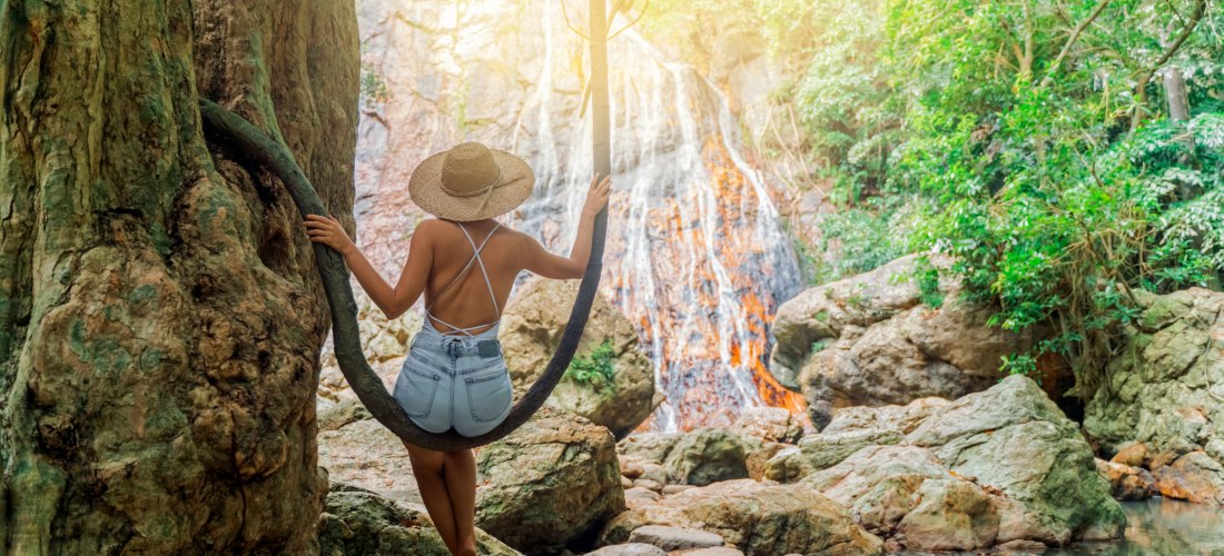 Das Bild zeigt eine junge Frau mit Strohhut und sommerlicher Kleidung, die barfuß auf einer natürlichen Lianenschaukel sitzt. Sie ist mit dem Rücken zur Kamera gewandt und blickt auf einen Wasserfall, der über moosbewachsene Felsen in ein kleines, von Ste