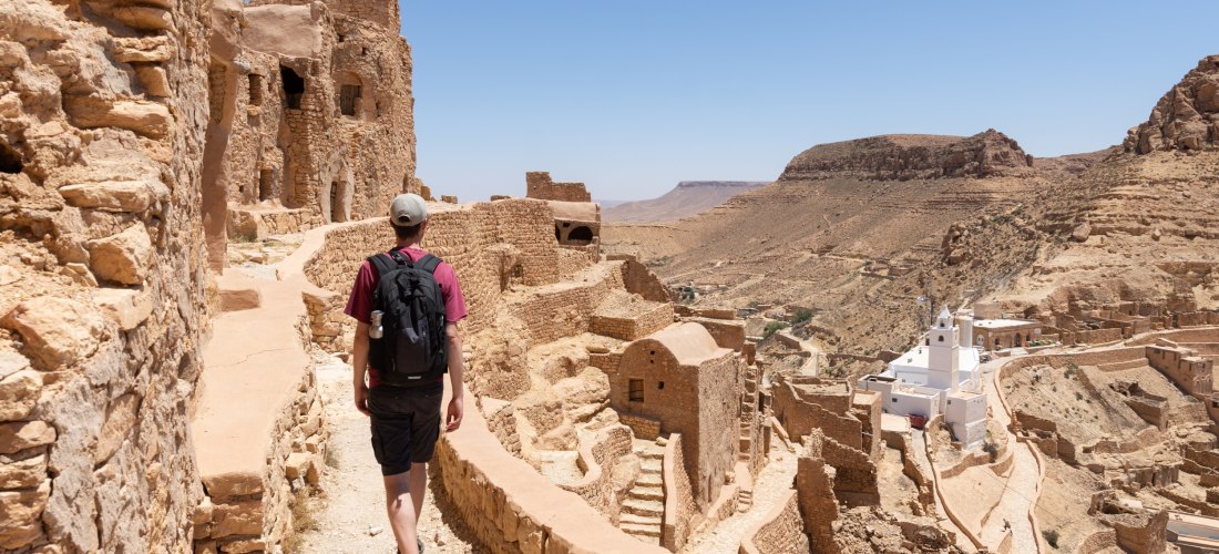 Ein Tourist mit Rucksack und Kappe geht auf einem schmalen Weg durch die Ruinen einer historischen Wüstenstadt aus Lehmstein. Rechts unten ist ein weißes Gebäude mit Türmen zu sehen, das sich von der sonst ockerfarbenen Landschaft abhebt. Im Hintergrund e