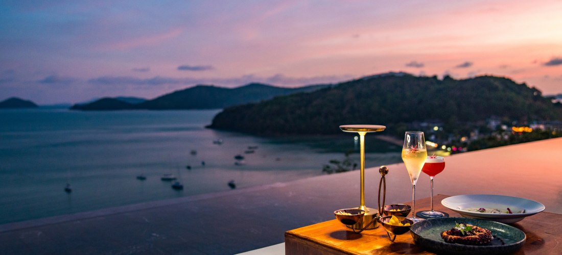 Abendstimmung über dem Meer auf Phuket. Im Vordergrund ein elegant gedeckter Tisch mit modernen goldenen Lampen, einem Glas Champagner, einem Cocktail sowie kunstvoll angerichteten Speisen. Im Hintergrund die Bucht und sanft beleuchtete Hügel unter einem 