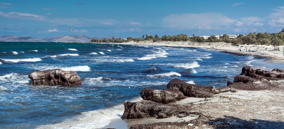 Langer Sandstrand von Mastichari auf Kos mit flachen Wellen, Felsbrocken und Seegrasanschwemmungen an der Nordküste.